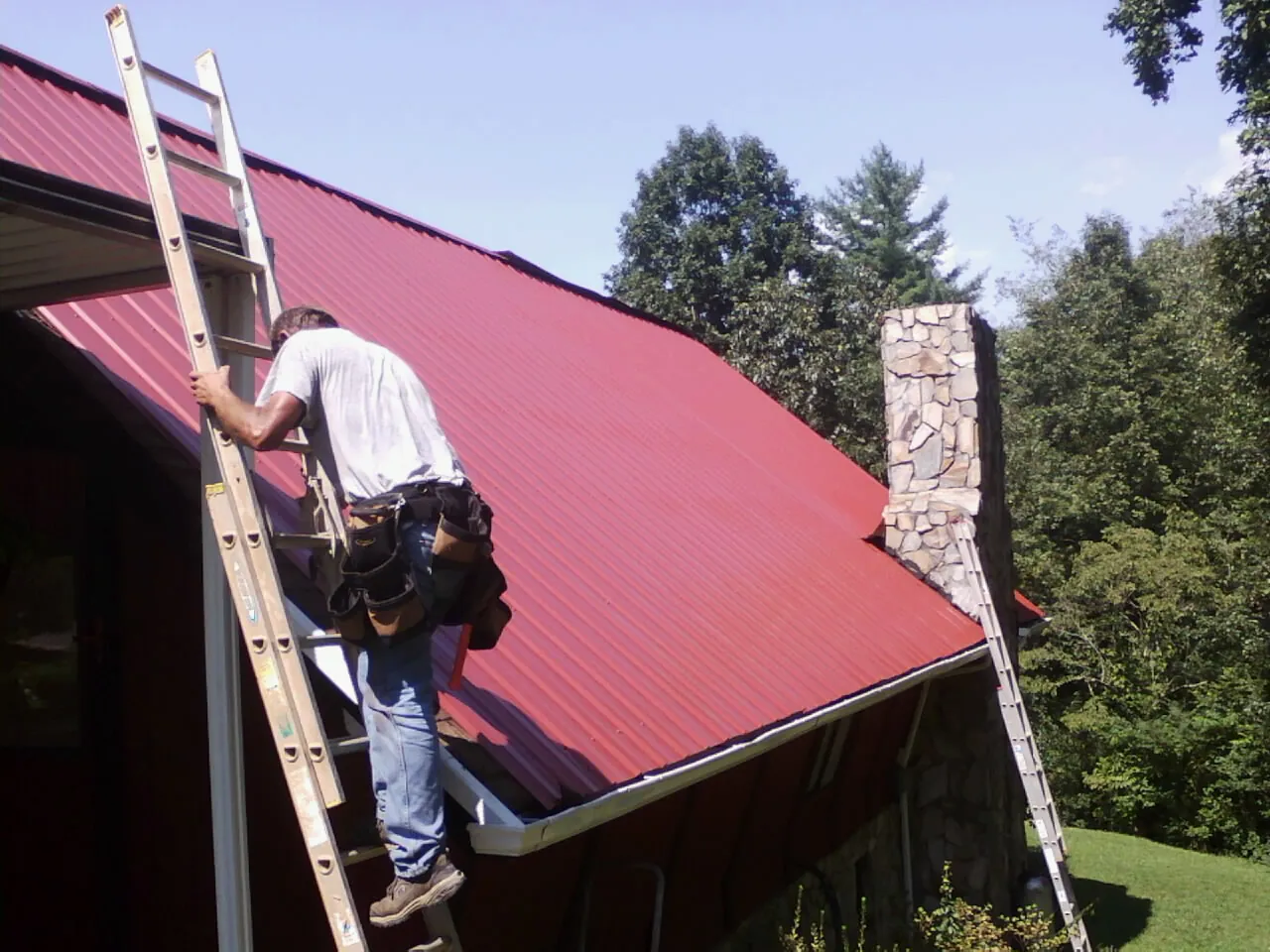 Roofer on a ladder beside a red metal roof and stone chimney — Soffit & Fascia Repair by Roofing Brothers in West Fork